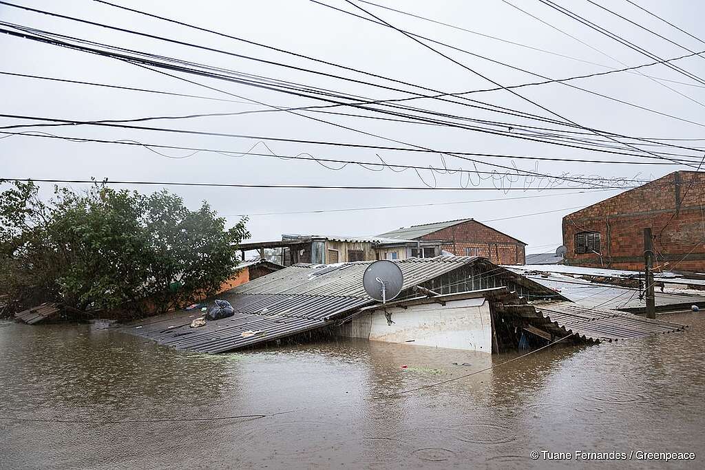 GP0SU0PPT_PressMedia_cprt_reduced A imagem mostra uma área alagada no bairro Rio Branco, em Canoas, Rio Grande do Sul, afetada por uma grave enchente ocorrida em 2024. A água cobre completamente a casa, mostrando somente um pedaço do telhado e do topo das moradias. No telhado visível, há objetos espalhados, incluindo um saco preto e destroços. A imagem é creditada a Tuane Fernandes.